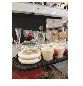 A tea set consisting of a transparent glass teapot with a black porcelain lid and a decorated red handle, four cream-colored porcelain cups with black and red spherical handles, and a rectangular blac
