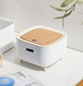 Small modern rectangular trash bin or storage box with rounded edges made of creamy white plastic with light beige legs and frame and light wooden lid with push button handle for easy opening 
