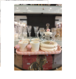 A tea set consisting of a transparent glass teapot with a light pink porcelain lid, four cream-colored porcelain cups with pink spherical handles, and a rectangular pink serving tray.